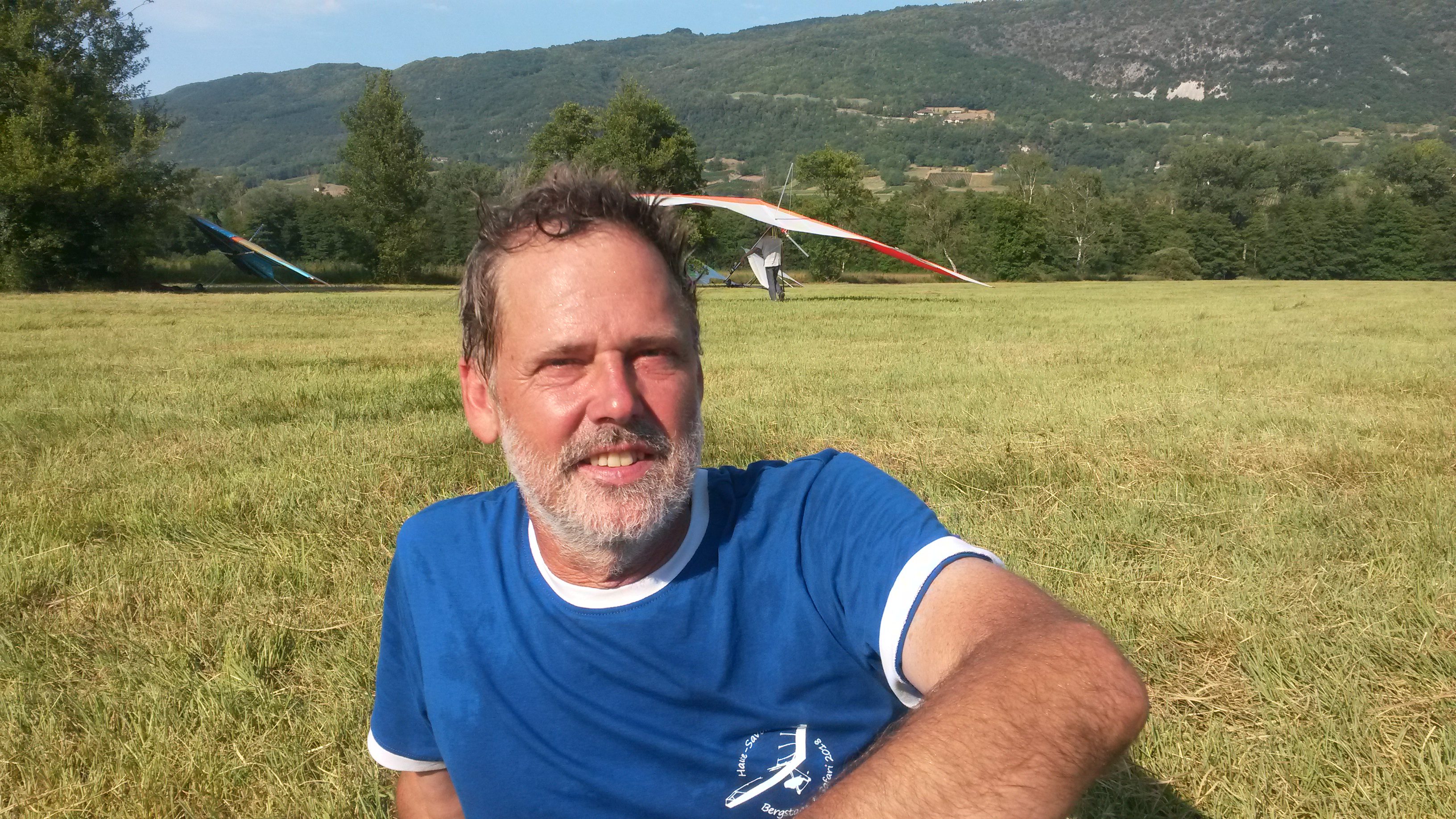 Foto van een man, zittend op een veld in de zon, met een landende deltavlieger op de achtergrond